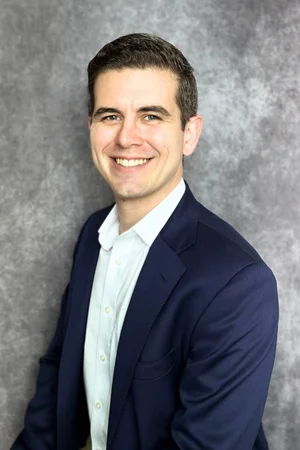 Smiling businessman in a navy suit against a gray background.